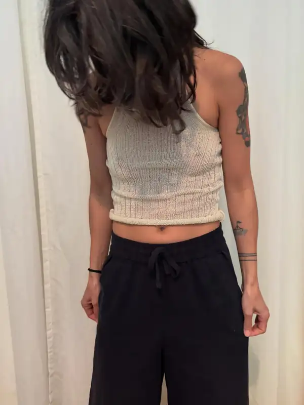 Torso: cream ribbed knit halter with a rolled hem, black trousers, hair falling forward, white curtain behind.