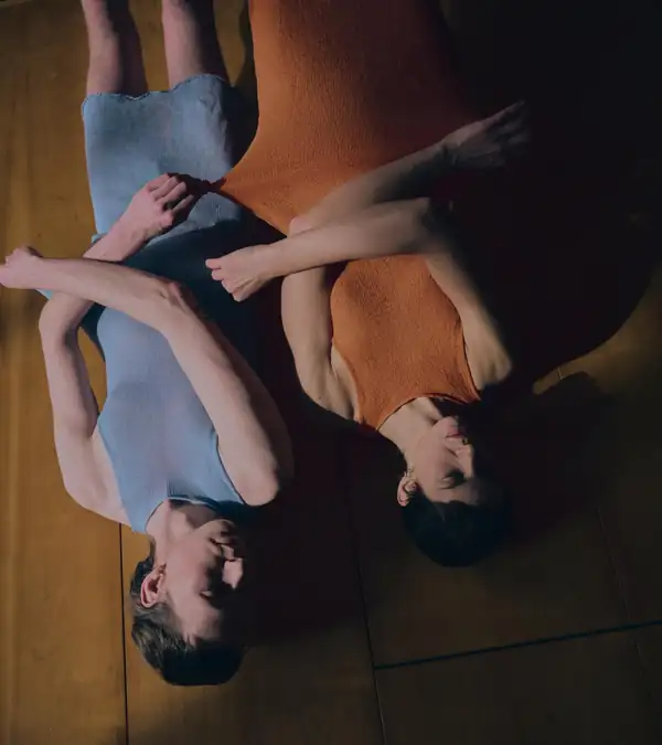 Overhead, rotated: two performers on a wooden floor, one in a pale blue knit dress, one in an orange knit dress.