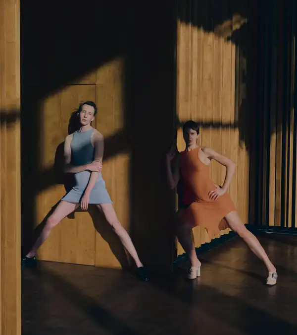 Two performers against a wooden wall in raking light, blue and orange knit dresses, legs braced.