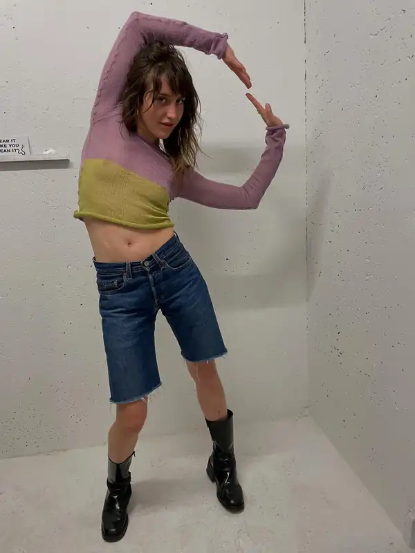 Full-length: lilac sheer long-sleeve over a chartreuse knit tube top, denim shorts, black boots, arms framing the head.
