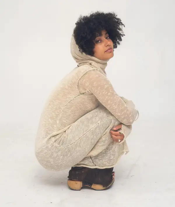 Crouching: cream flecked knit long dress with a hood/cowl and long sleeves, brown clogs.