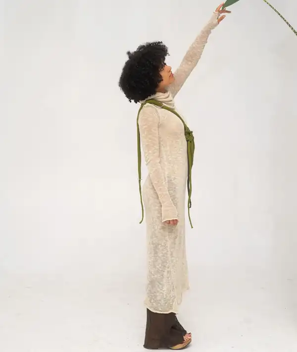 Full-length, arm raised to a leaf: the cream long dress and olive bag.