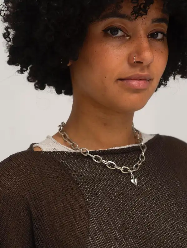 Close portrait: brown sheer knit top, chunky silver chain with a heart.