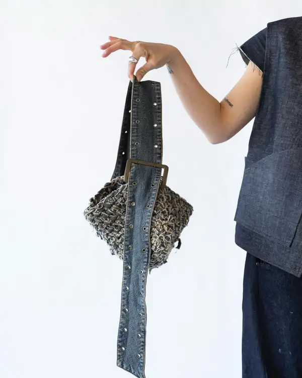 A hand holding a knitted bag on a denim strap with a wooden handle.