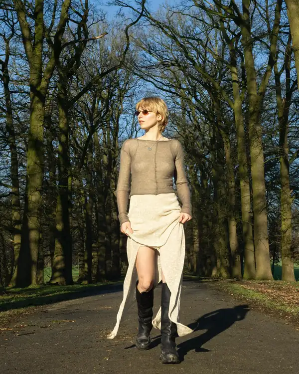 Full-length in a bare-tree avenue: sheer taupe knit top over a long cream knit skirt held wide, black knee boots. IDENTICAL to 24-03.