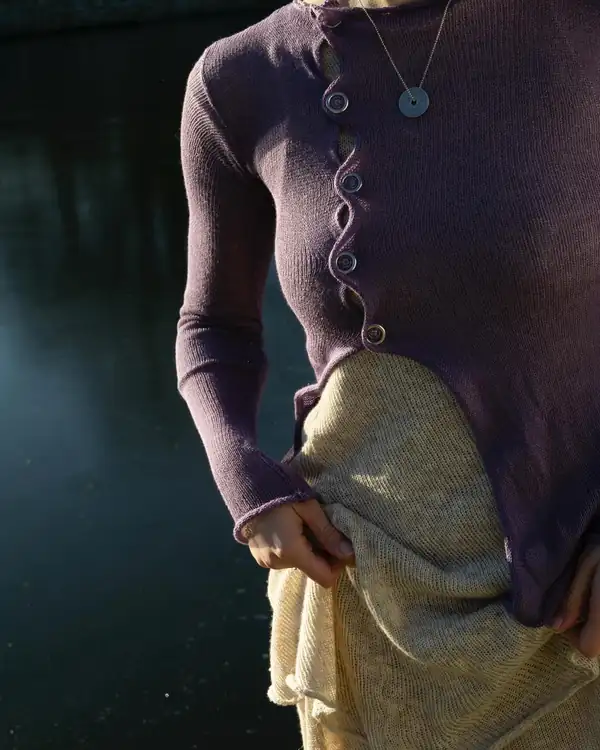 Close: purple knit asymmetric cardigan with a row of shell buttons, over a cream knit skirt, dark water behind.