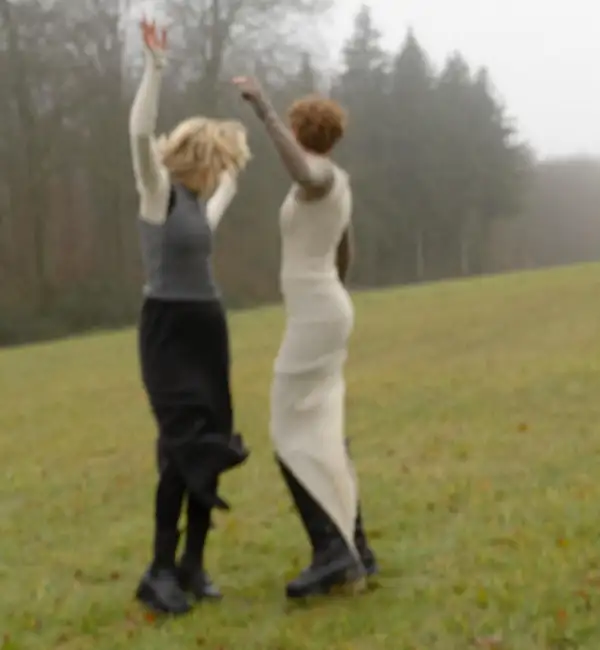 Two models from behind in a misty field, arms raised, grey and cream knit long dresses.