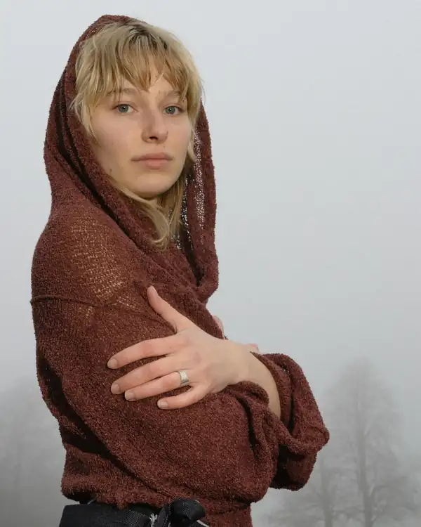 Portrait: the rust knit hood wrapped around the head and shoulders, arms folded.