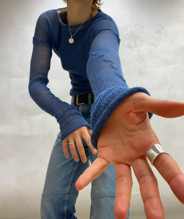 Leaning: blue sheer knit long-sleeve, jeans, a hand thrust to camera with a wide silver ring.