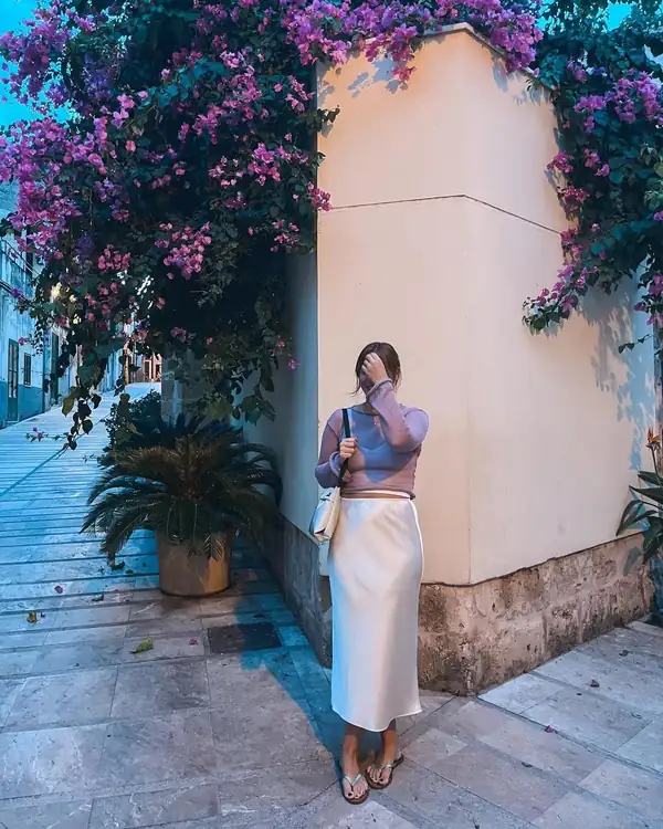 Full-length by a pink wall under bougainvillea: lilac sheer knit long-sleeve over a white satin skirt.