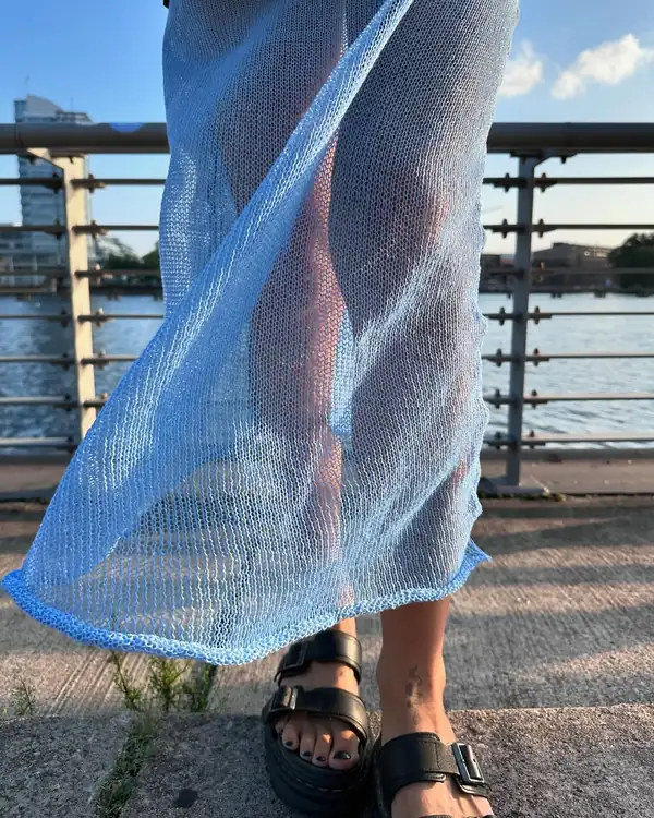 Pale blue sheer knit long skirt caught by the wind across the leg, black sandals, river behind.