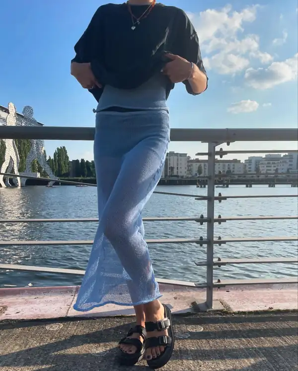 Adjusting the black tee over the pale blue skirt, waterfront behind.