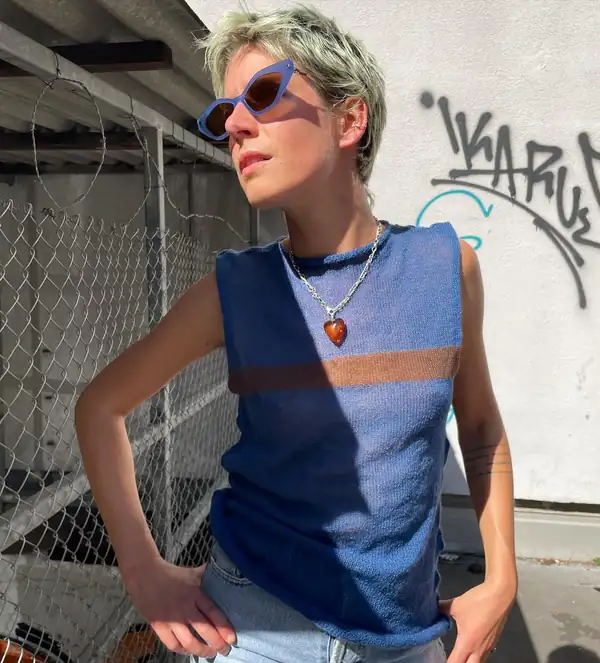 Navy and cornflower colour-blocked sleeveless knit with a rust stripe, denim shorts, blue cat-eye sunglasses, graffiti behind.