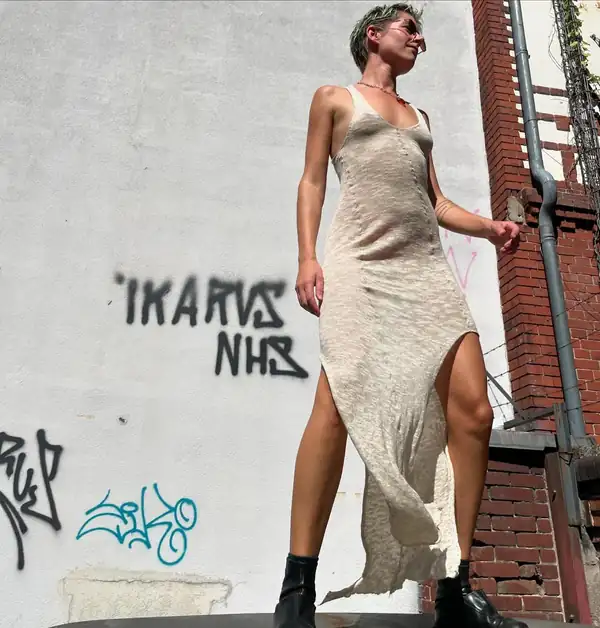 Full-length: cream/oat knit long dress with a high side slit, black boots, graffitied wall.