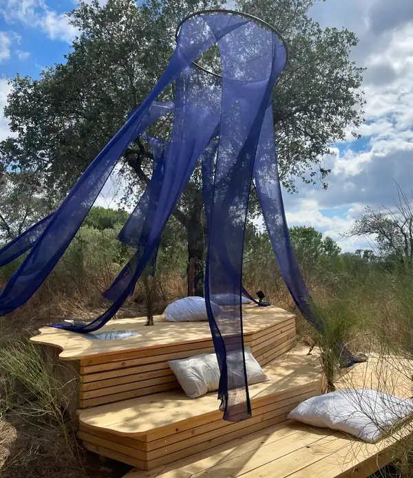 An installation of long navy sheer knitted panels hanging from a ring over a wooden deck with cushions, trees and sky behind.