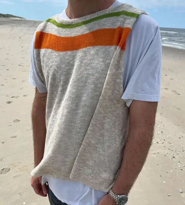 A man in an oatmeal knit sleeveless tabard with green and orange chest stripes over a white tee, beach behind.