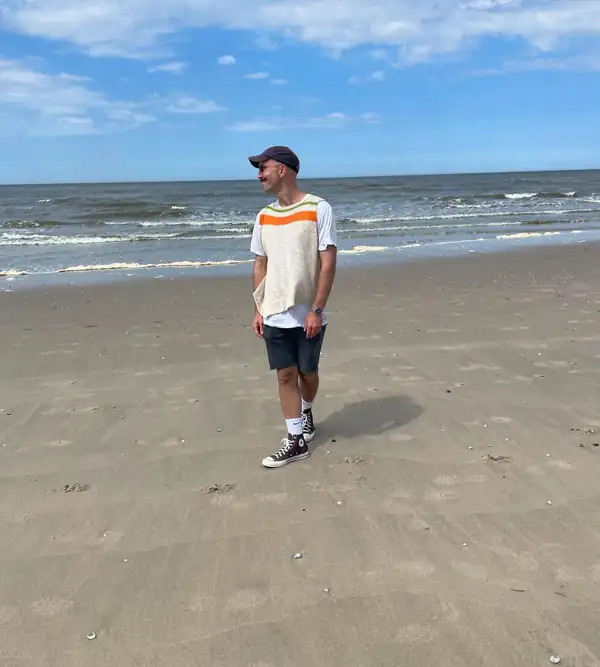 Full-length walking on the beach: the striped vest over a white tee, denim shorts, cap.