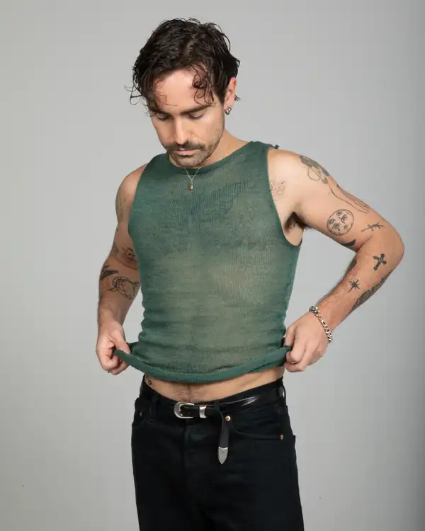 A man pulling the hem of a sage-green sheer knit tank straight, black jeans.