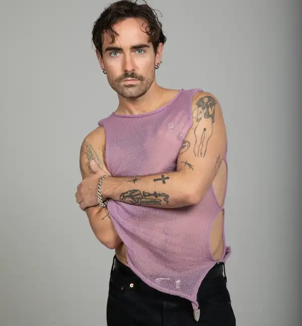 A man in a lilac knit sleeveless top with open, unseamed sides, arms crossed.