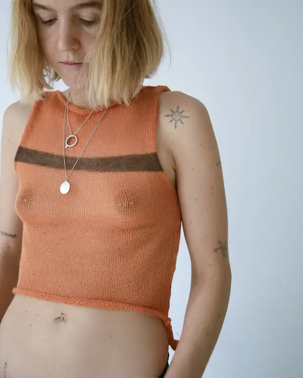 Orange knit sleeveless top with a brown chest stripe, silver pendants, black trousers. Nipples visible through the knit.