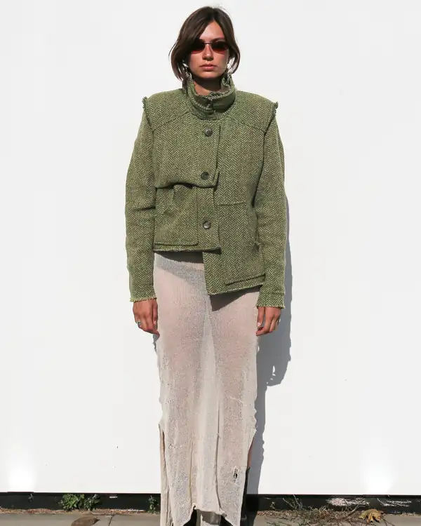 Full-length: olive herringbone knit jacket with a high funnel collar over a long cream sheer knit skirt, black boots.