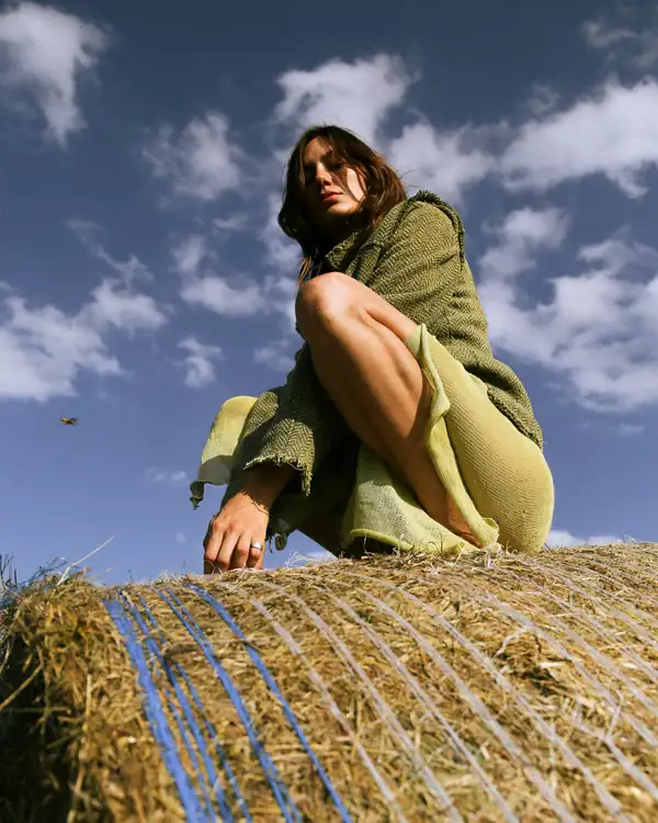 Crouched on a round hay bale against a blue cloudy sky: olive knit jacket over a chartreuse knit dress.
