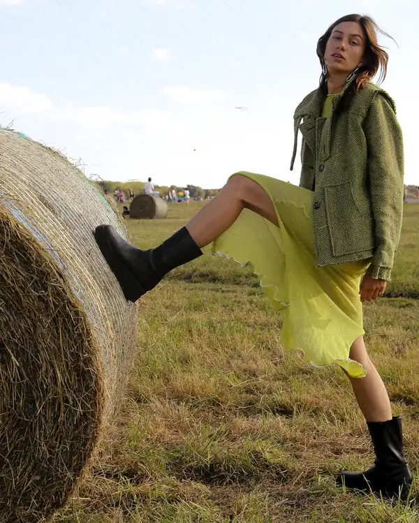 Leg braced against the bale: the olive jacket over the chartreuse sheer knit dress, black boots.