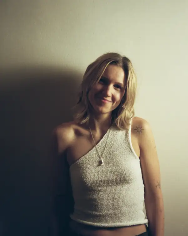 Half-length portrait of a smiling person with shoulder-length blond hair, in a white textured one-shoulder knit top and a pendant on a chain, against a plain pale wall.